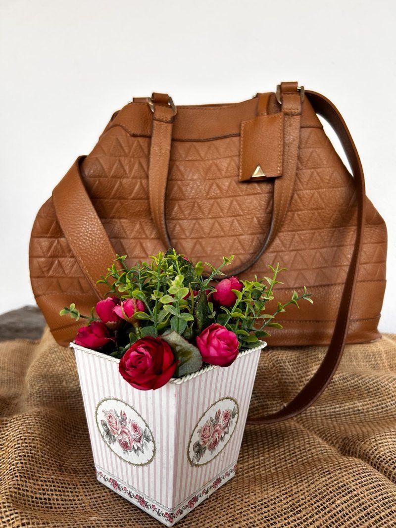 Bolsa Schütz em couro caramelo tamanho G, com alça transversal, apoiada sobre juta ao lado de um vaso com flores